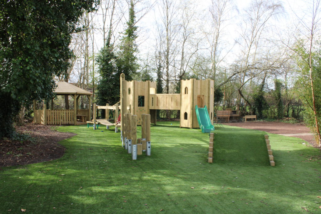 Outdoor playground at Rufford Primary School featuring wooden climbing structures, a slide, rope bridge, gazebo, and artificial grass, all surrounded by trees and greenery for a fun and safe play environment.