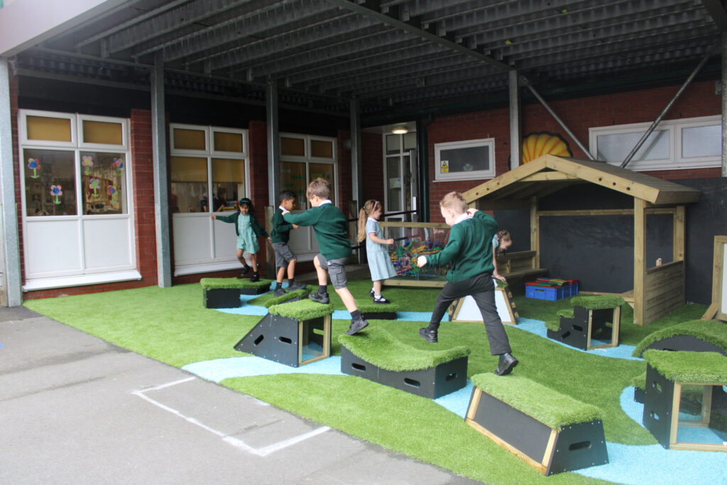 Several children play on artificial grass and plastic stepping platforms outside St Kentigern's school building, with a small wooden playhouse in the background.