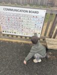 A young child crouches and points at a large Playground Communication Board mounted on a fence in a playground area.