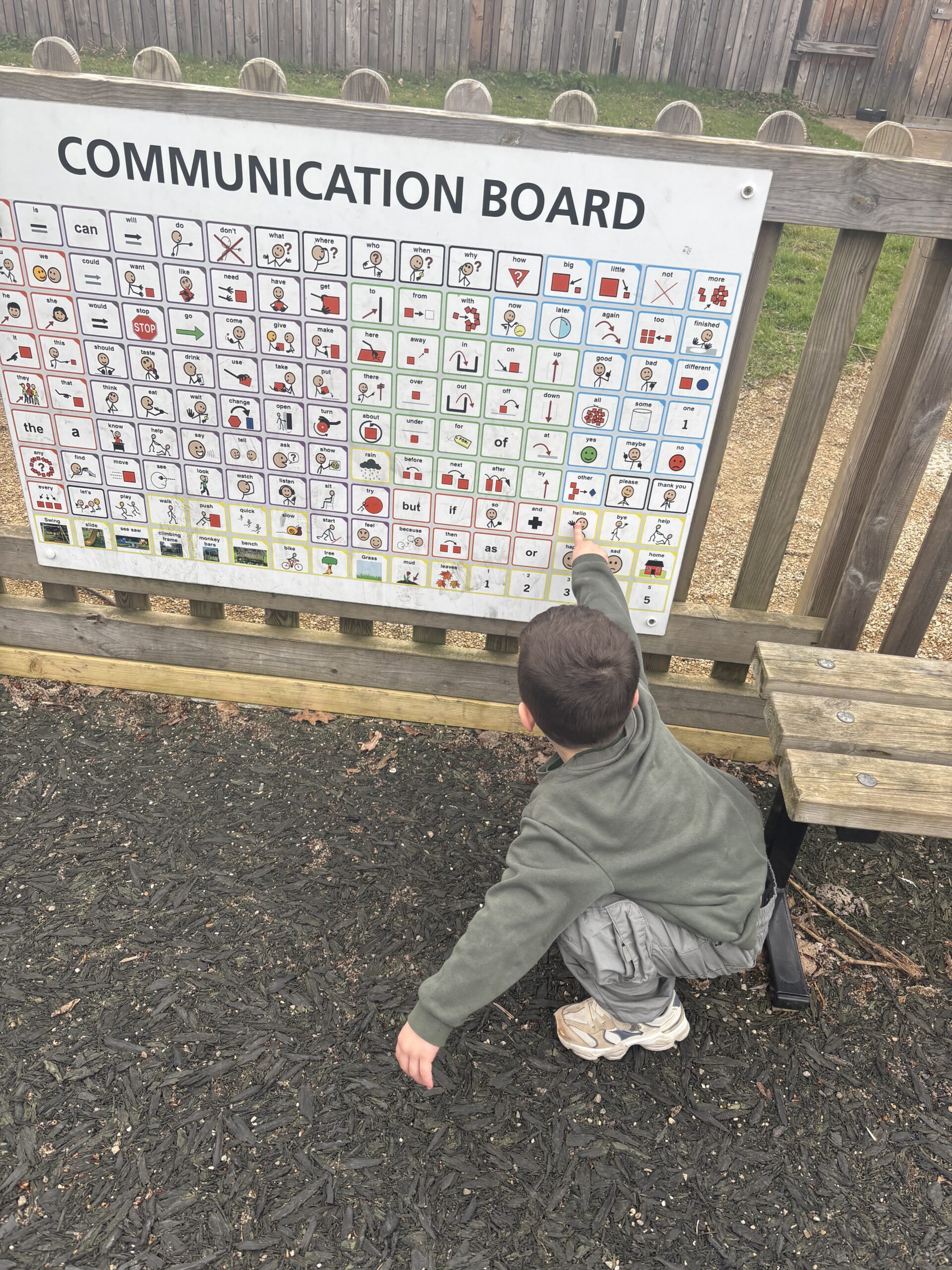 A young child crouches and points at a large Playground Communication Board mounted on a fence in a playground area.