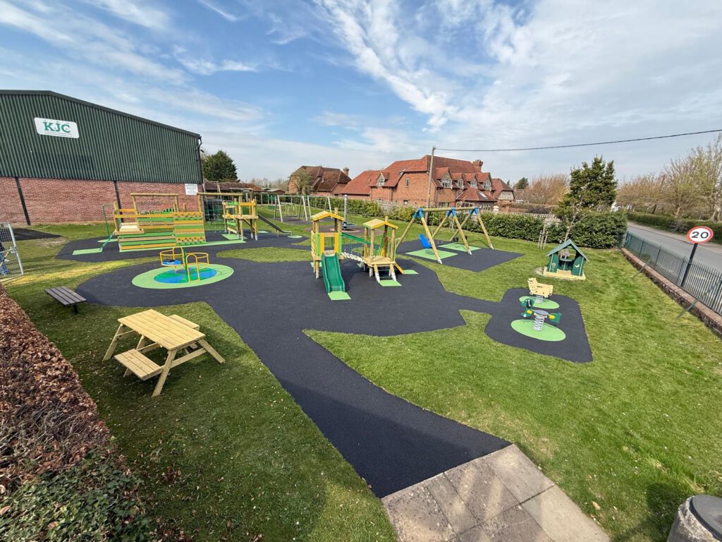 Outdoor playground with green and yellow equipment, picnic benches, and a small playhouse on grassy black safety flooring—maintained by Kintbury Parish Council—located near a road and brick buildings.