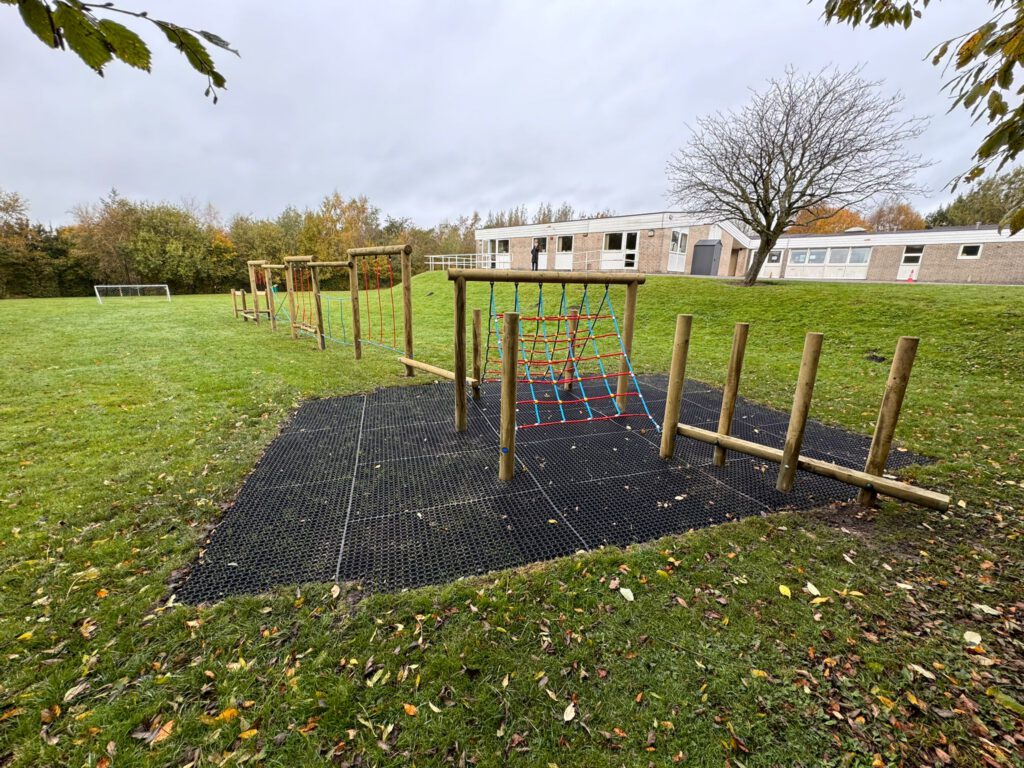 Outdoor playground with wooden climbing structures and a rope net on a grassy field. Buildings and trees are in the background.
