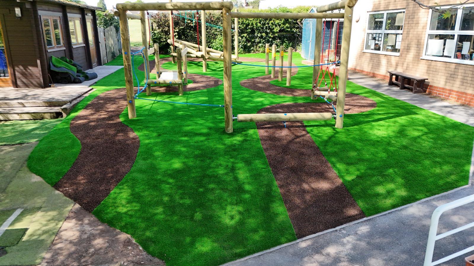 Outdoor playground with artificial grass, a winding brown path, and wooden climbing structures next to a building with windows and benches.