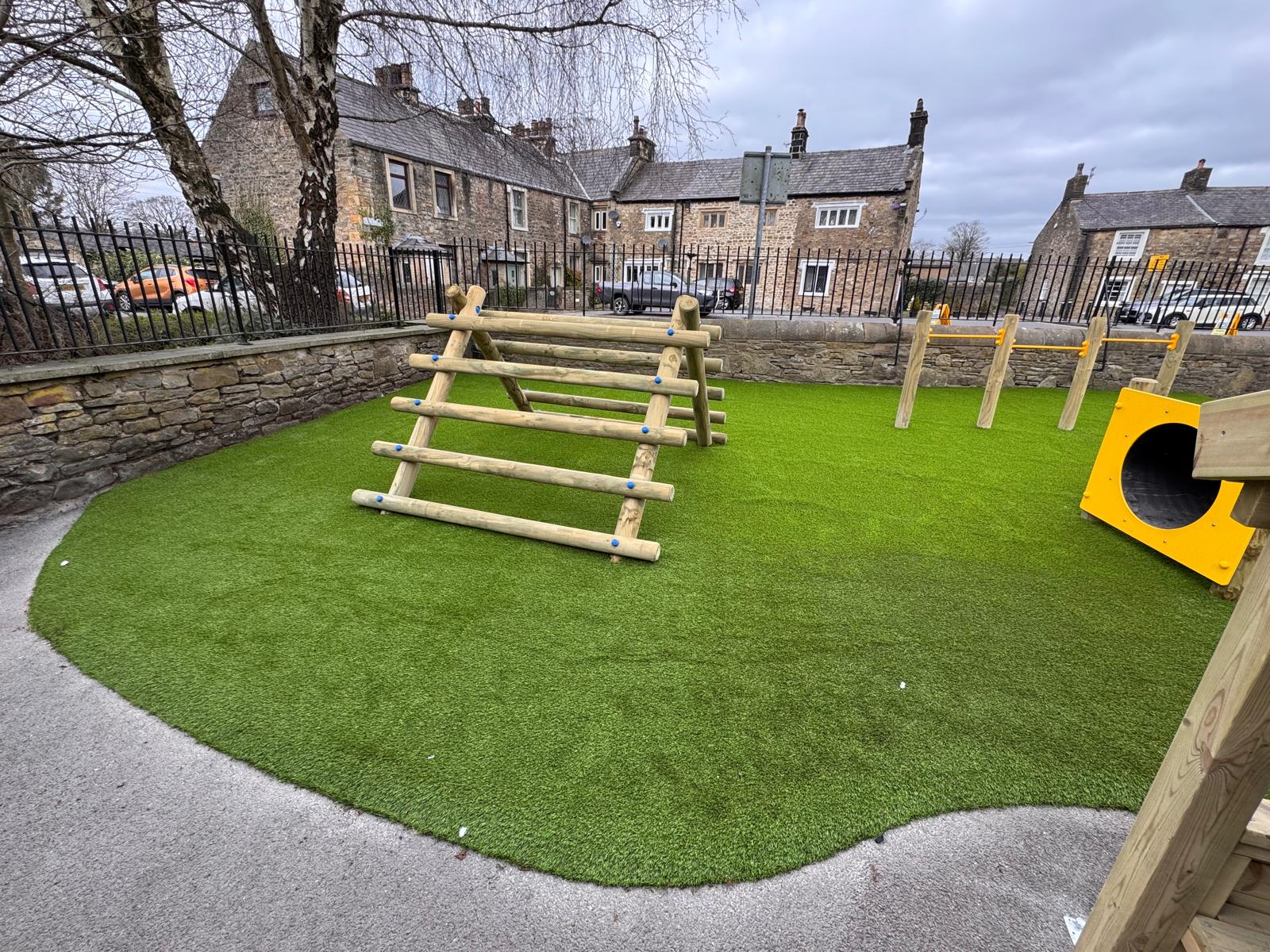 A small playground with artificial grass features a wooden climbing frame, yellow tunnel, and wooden posts, surrounded by a stone wall and residential houses in the background.