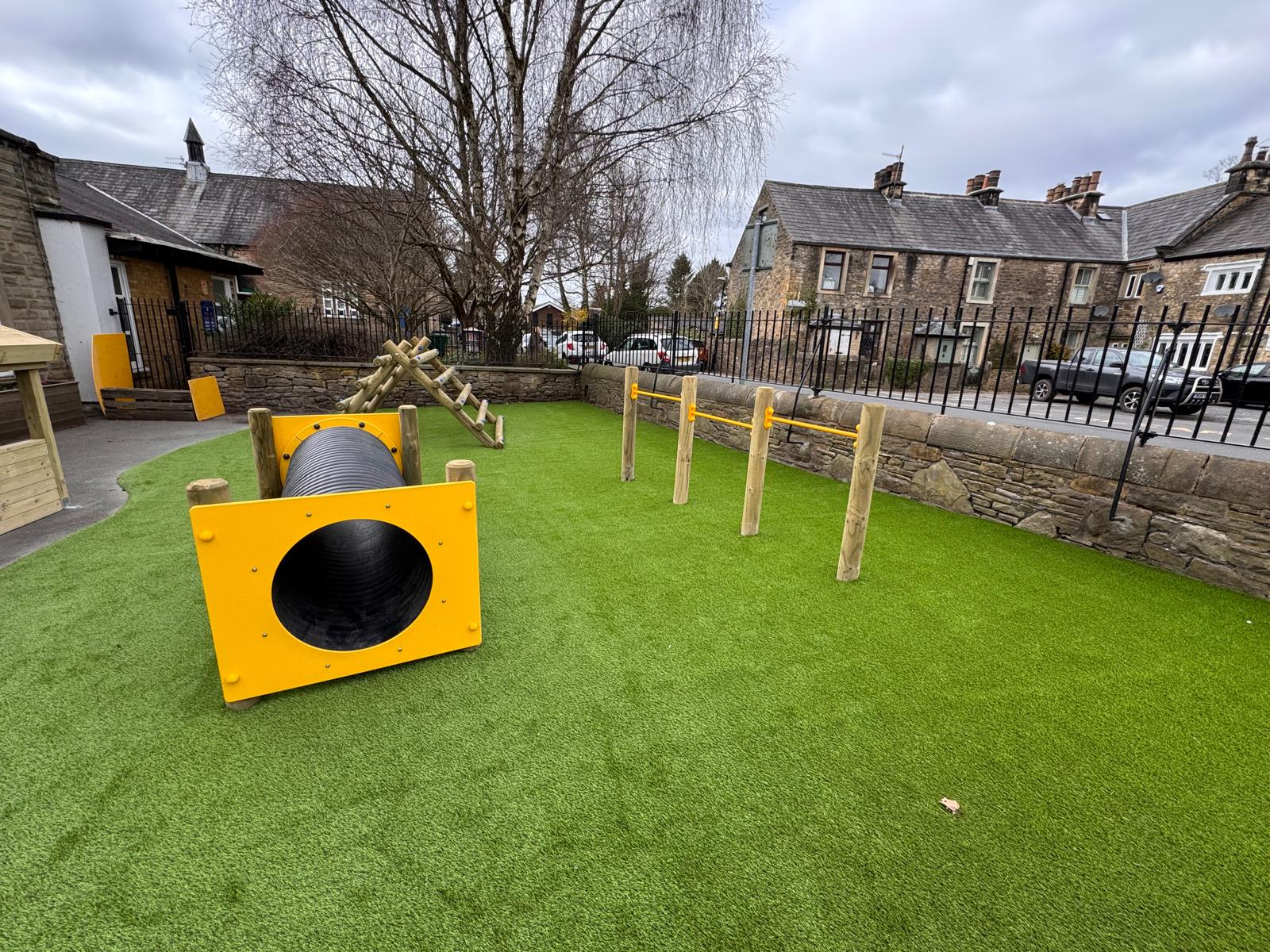 Childrens outdoor play area with artificial grass, a black tunnel with yellow ends, climbing bars, and wood structures, surrounded by a stone wall and metal fence near residential buildings.