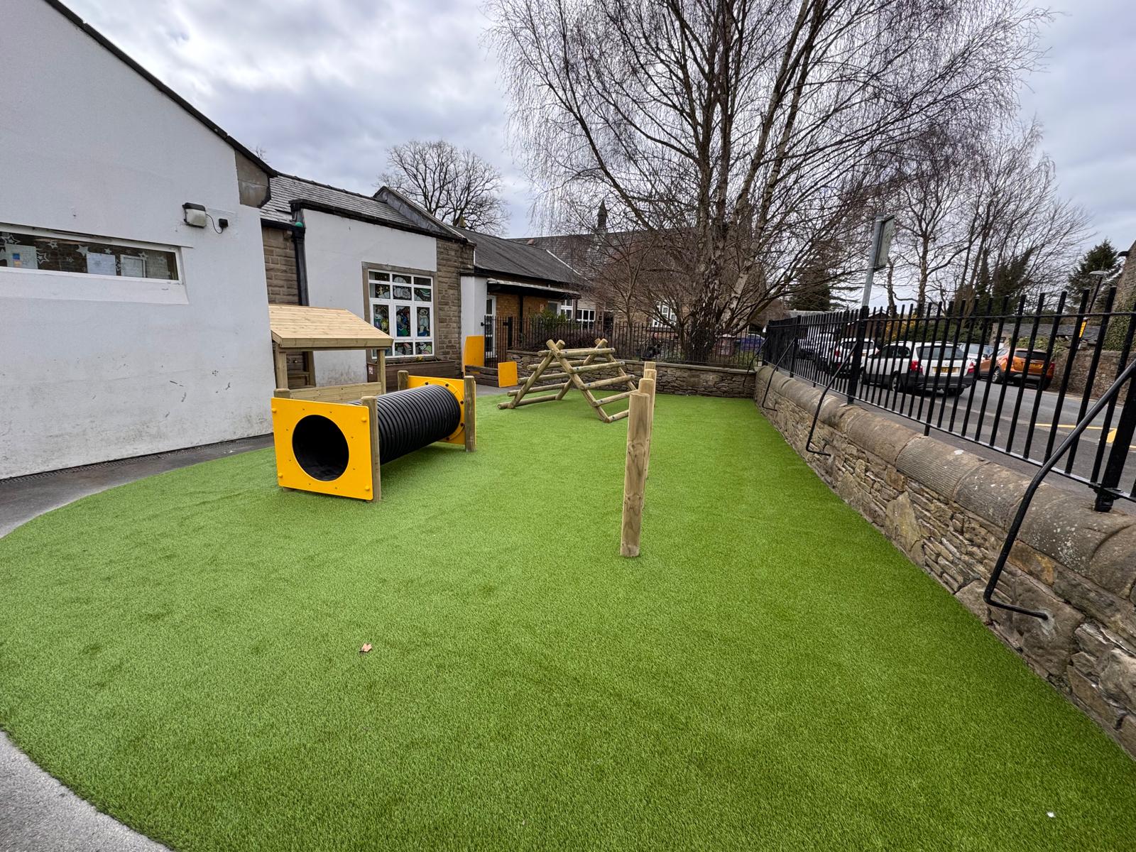 Outdoor playground with artificial grass, wooden climbing structures, a yellow tunnel, and nearby buildings and a fence under cloudy sky.