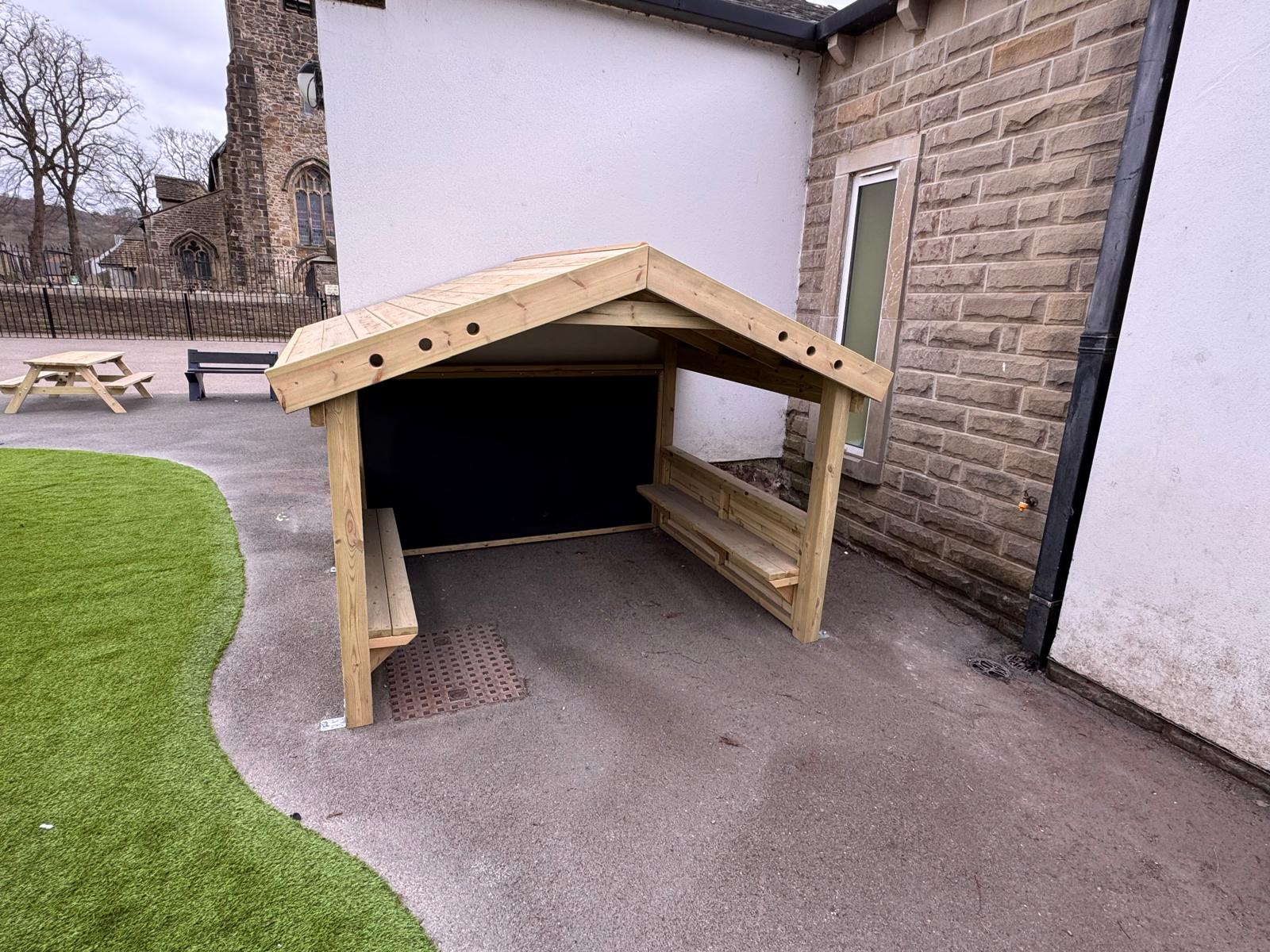 A small wooden shelter with benches and a slanted roof stands next to a building on a paved outdoor area near some artificial grass.