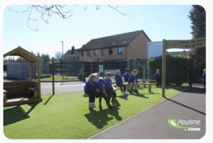 A group of children in blue uniforms sit and talk on benches in a fenced school playground with artificial grass on a sunny day.
