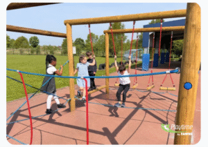 Four children play and climb on a wooden rope playground structure outdoors on a sunny day, with grass and trees in the background.