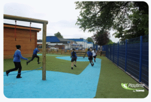 Four children in school uniforms run and play on a playground with artificial grass and blue markings, near a wooden structure, fencing, and trees. The logo Playtime by FAWNS is visible.