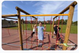 Three children play on a wooden climbing frame with ropes at a playground under a blue sky. The girl in front smiles and holds onto the ropes.