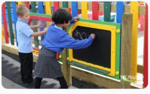 Two children in school uniforms draw on an outdoor chalkboard attached to a colorful play fence.