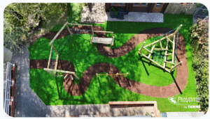 Aerial view of a playground with wooden climbing structures and swings on artificial grass and winding brown paths.