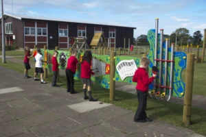 Six children in red and black school uniforms play on outdoor musical instruments in a school playground area on a clear day.