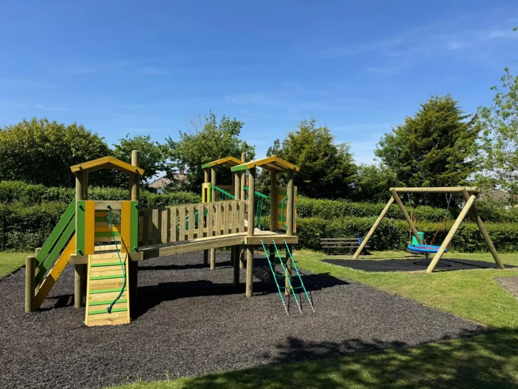 A wooden playground structure with slides, ladders, and a bridge stands on a rubber safety surface next to a swing set with a large blue disc swing, surrounded by grass and trees.