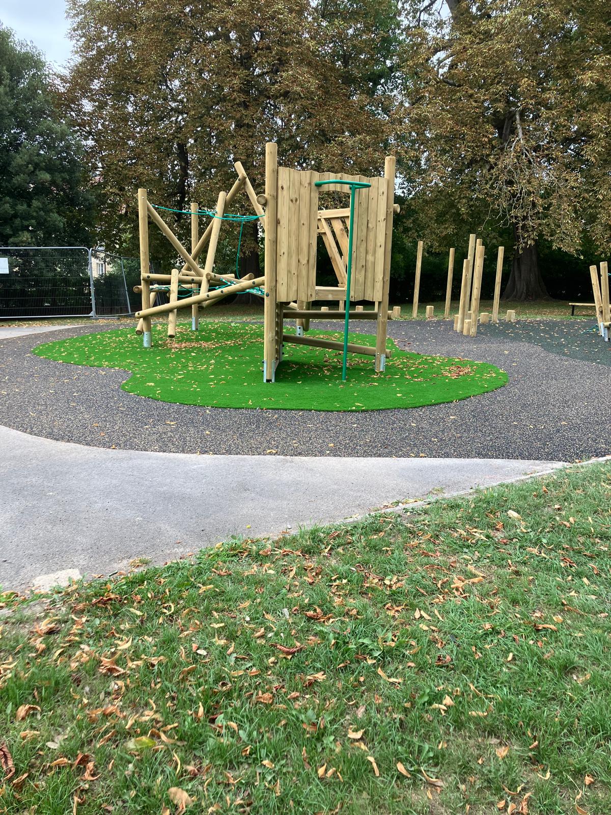A wooden playground climbing frame stands on artificial grass, surrounded by a path and trees with fallen autumn leaves.