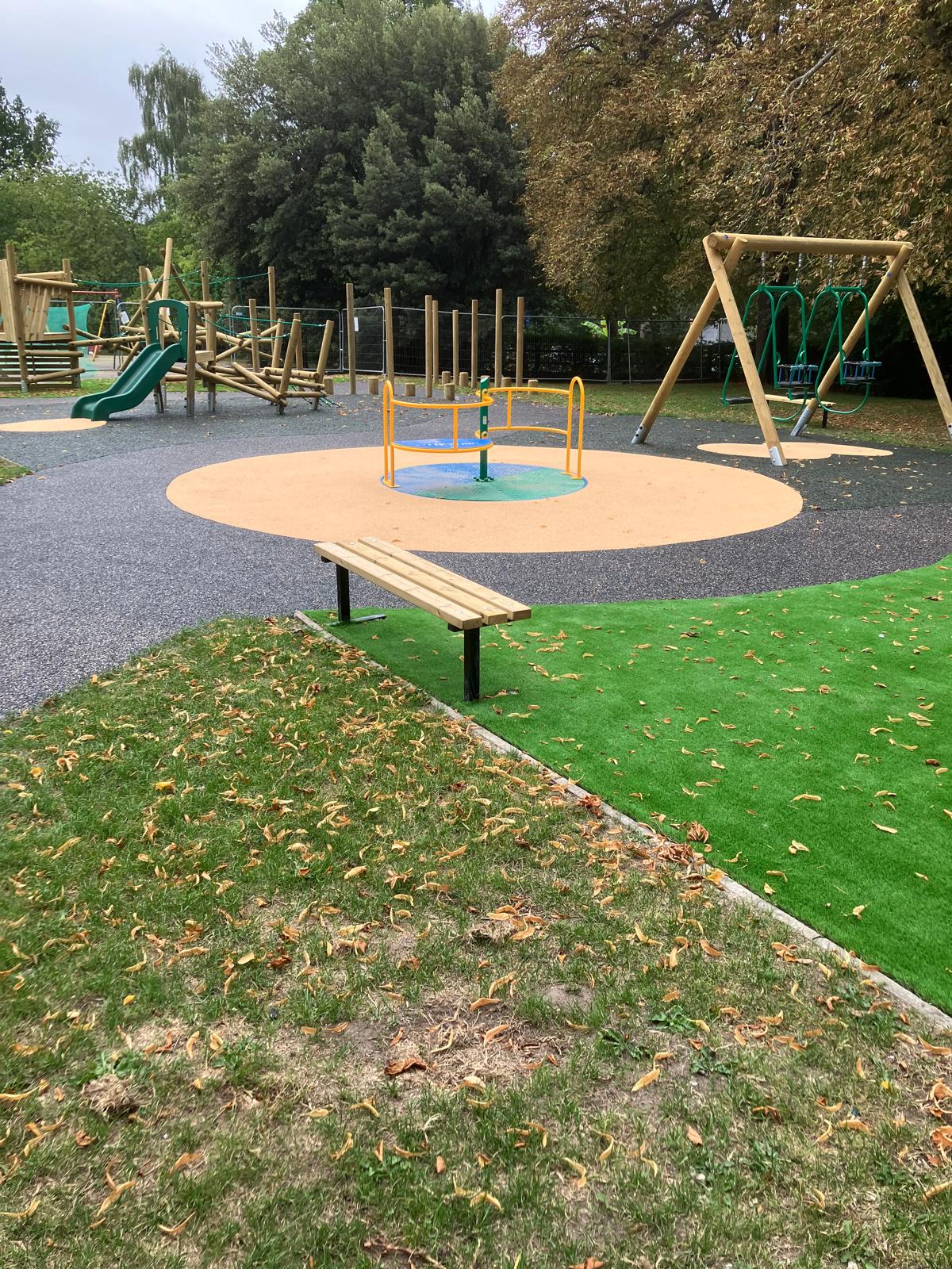 Outdoor playground with a roundabout, swings, slide, and climbing frames, surrounded by grass and trees. Fallen leaves are scattered on the ground.