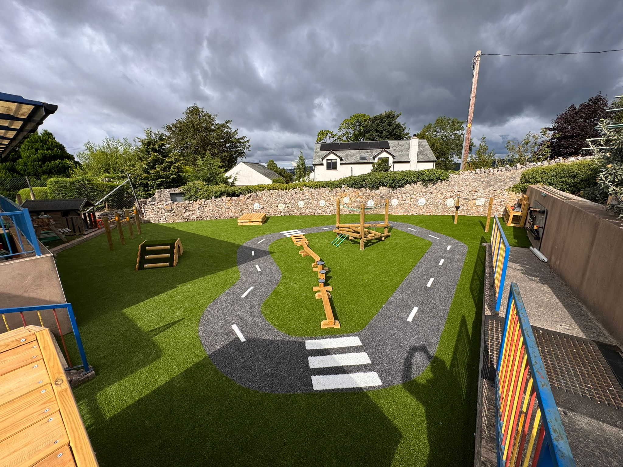 Outdoor playground with artificial grass, a painted road track, wooden climbing structures, and a stone wall in the background under a cloudy sky.