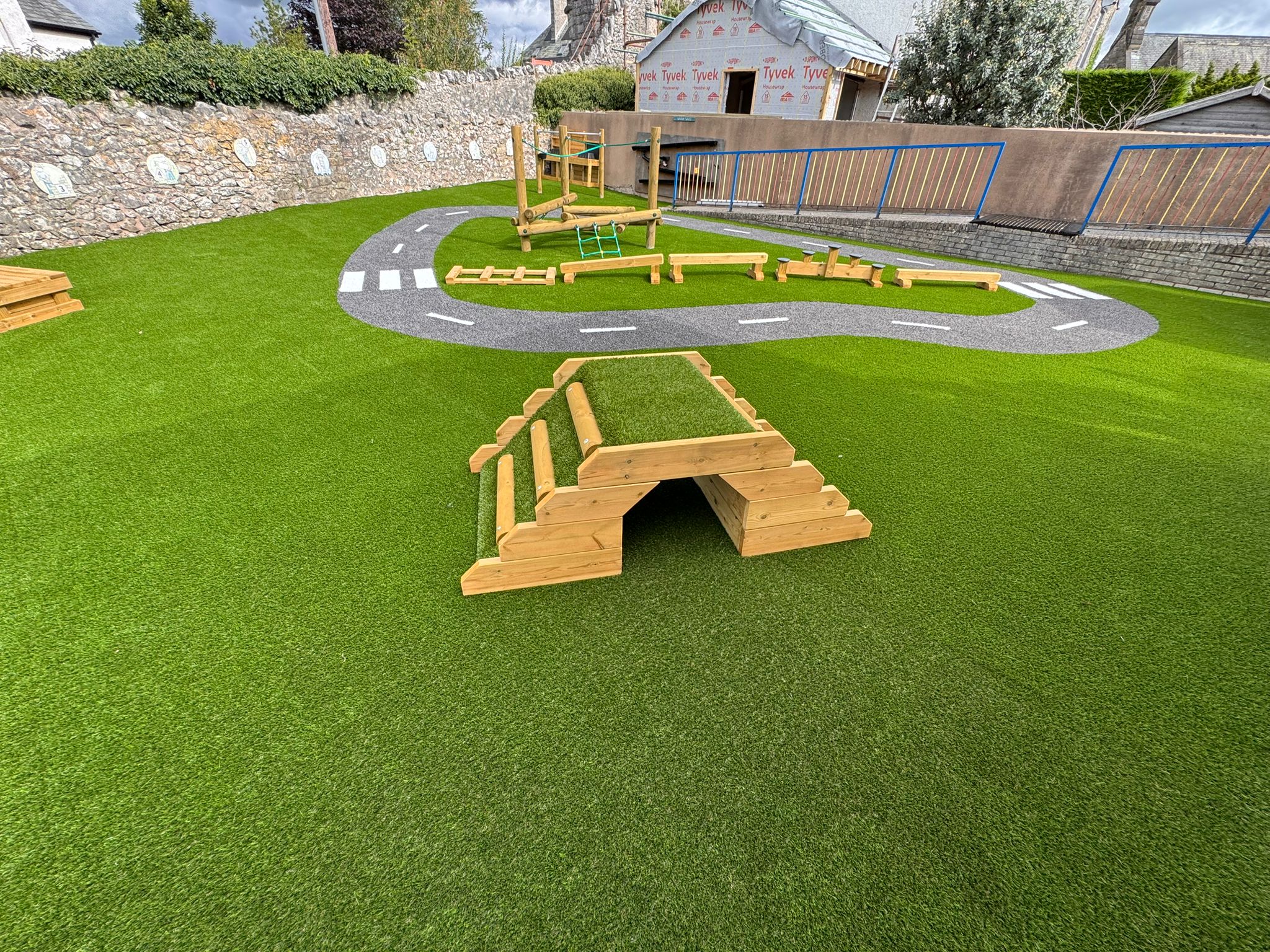 A playground with artificial grass, wooden climbing structures, and a winding bike or scooter path painted to look like a road.