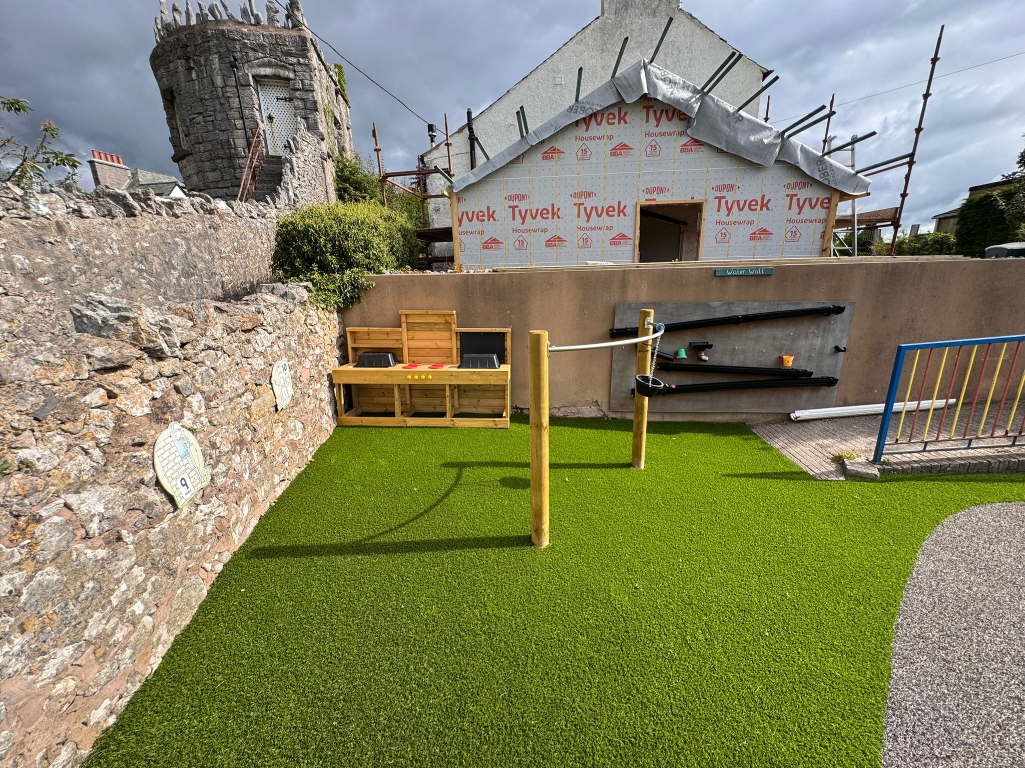 A small outdoor play area with artificial grass, wooden play structures, a water play set-up, and a building under construction in the background.