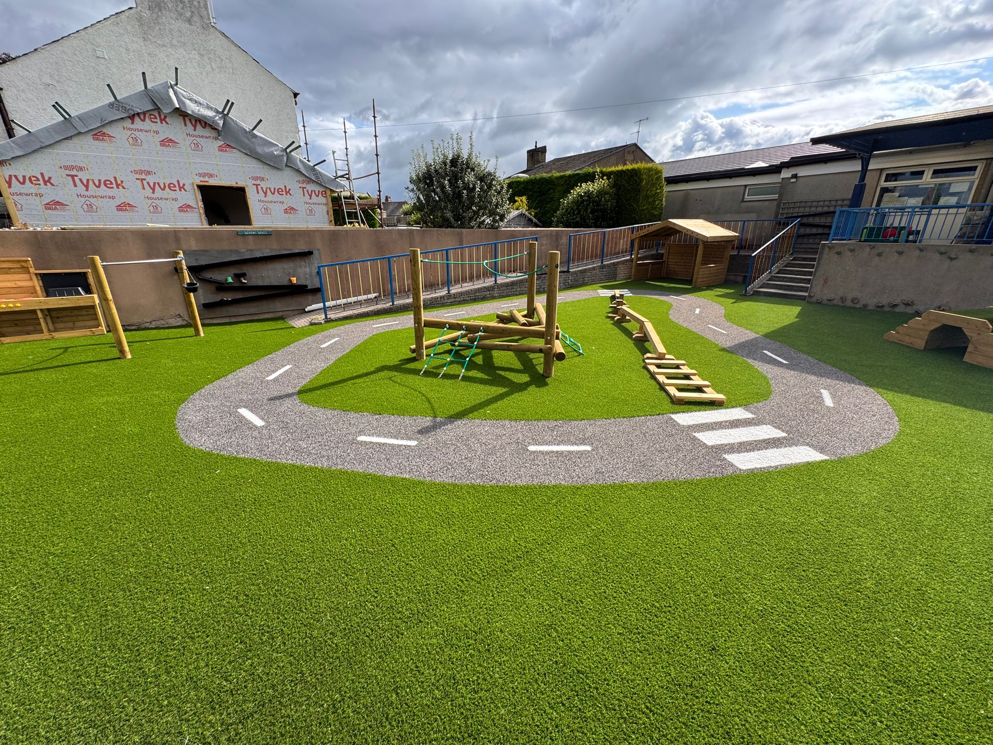 Outdoor playground with artificial grass, a painted road track, wooden climbing frames, and sheds, surrounded by buildings and a cloudy sky.