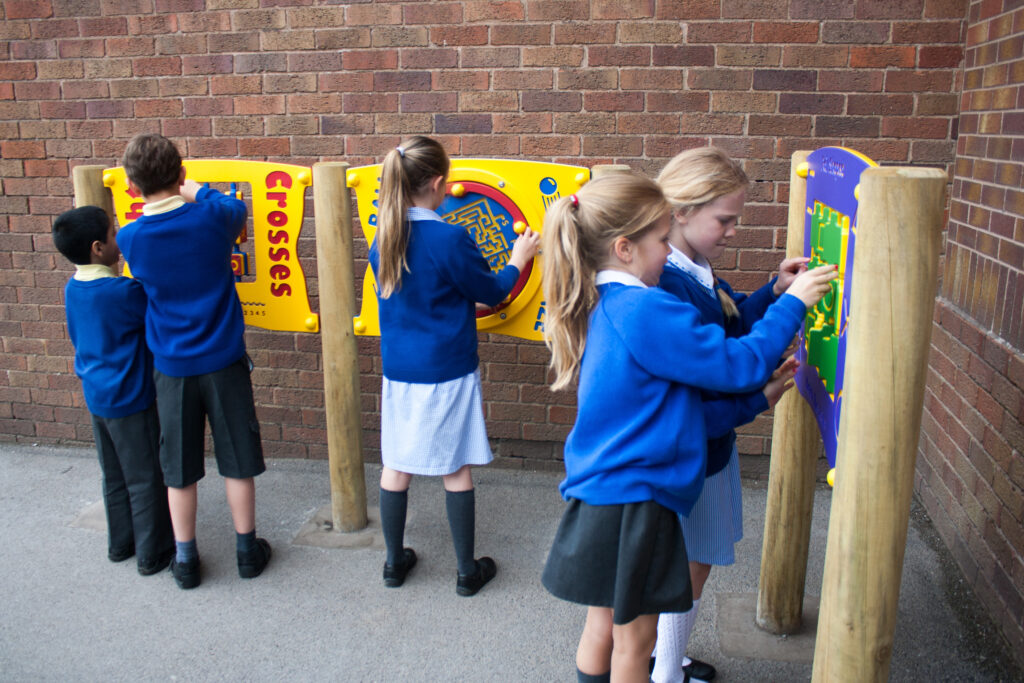 Five schoolchildren in uniform play with interactive wall panels mounted outdoors against a brick wall.