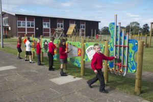 Six children in red school uniforms play with colourful musical panels on an outdoor playground, with a school building visible in the background.