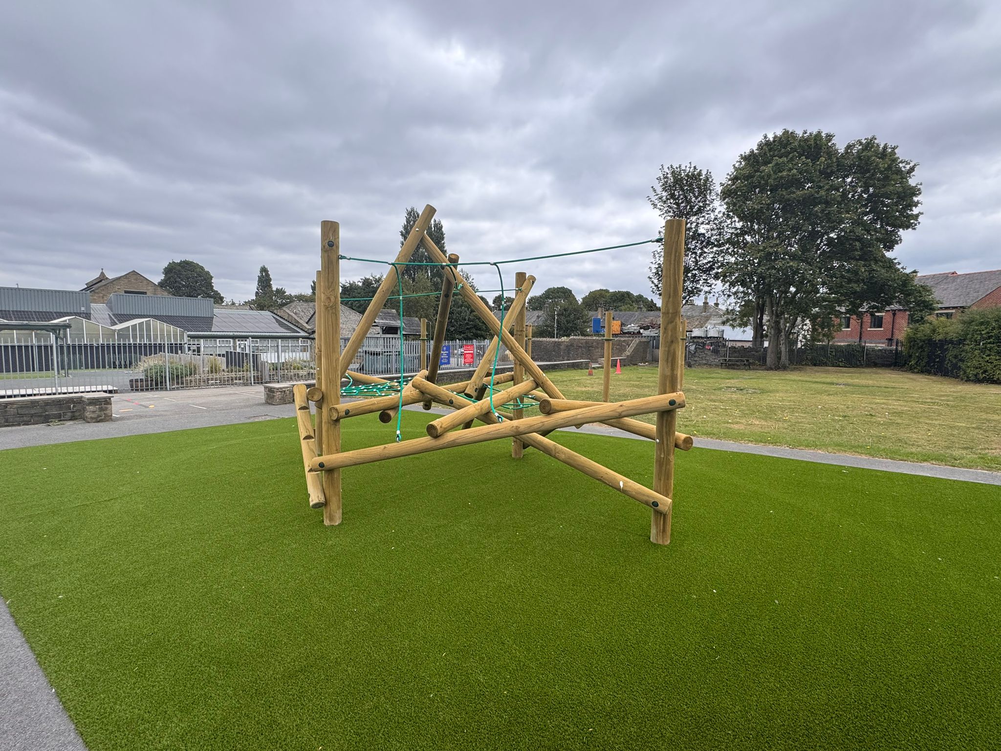 A wooden climbing frame with ropes is installed on artificial grass in an outdoor play area under a cloudy sky.