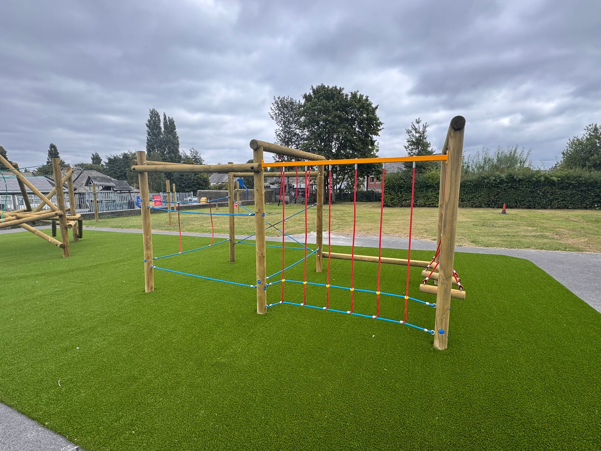 Children’s playground climbing frame made of wood and coloured ropes, situated on artificial grass under a cloudy sky. Paths and grassy areas surround the play equipment.