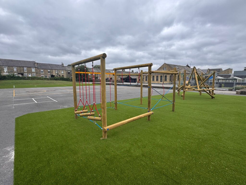 A playground with wooden climbing structures on artificial grass, next to a paved area with painted court lines. Stone buildings are visible in the background under a cloudy sky.