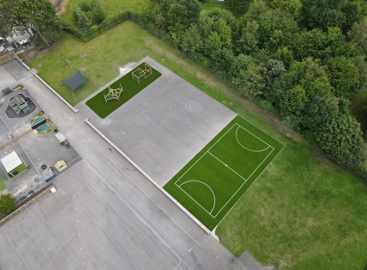 Aerial view of a playground with a basketball court, climbing frames, green grass areas, and surrounding trees.