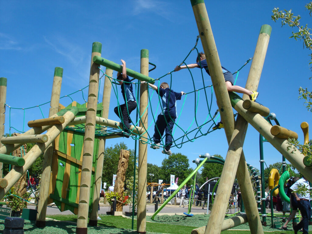 Three children climb on a wooden and rope playground structure outdoors under a blue sky, with more people and playground equipment in the background.