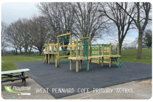 A wooden playground climbing structure with green accents sits on a black rubber surface at West Pennard CofE Primary School—an inspiring example of Playground Ideas for Primary Schools, with benches and grassy areas nearby.