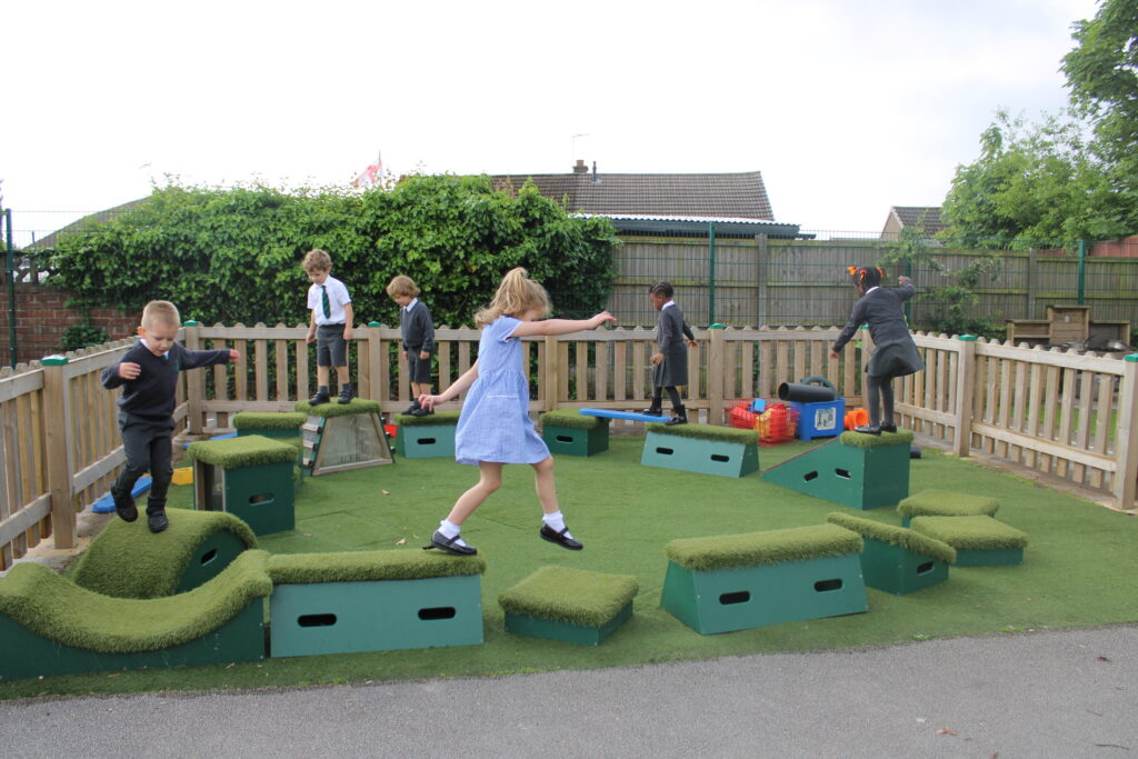 Several young children in school uniforms play and balance on artificial grass blocks in a fenced outdoor play area.
