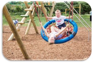 Two children are playing on a large rope swing in a playground with woodchip ground cover and climbing frames in the background.