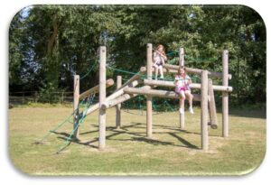 Two children sit on a wooden climbing frame with green ropes in an outdoor playground, surrounded by grass and trees.