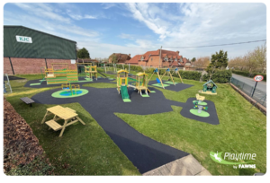 Outdoor playground with swings, slides, climbing frames, benches, and play equipment on a grass and rubber surface next to a green building and houses under a partly cloudy sky.