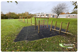 A school playground with wooden climbing apparatus, rope netting, and safety mats on grass; buildings and a football goal are visible in the background.