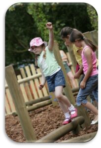 Three young girls balance and walk on wooden beams at an outdoor playground, with one girl raising her arm for balance.