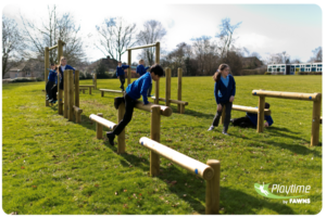 Children in school uniforms play on a wooden outdoor assault course in a grassy field under a partly cloudy sky. Playtime by Fawns logo is visible in the bottom right corner.