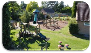 A playground with climbing frames, slides, swings, and play structures on a grassy area, surrounded by trees and buildings.