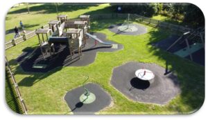 Aerial view of a fenced playground with climbing frames, slides, a roundabout, swings, and safety matting surrounded by grass.