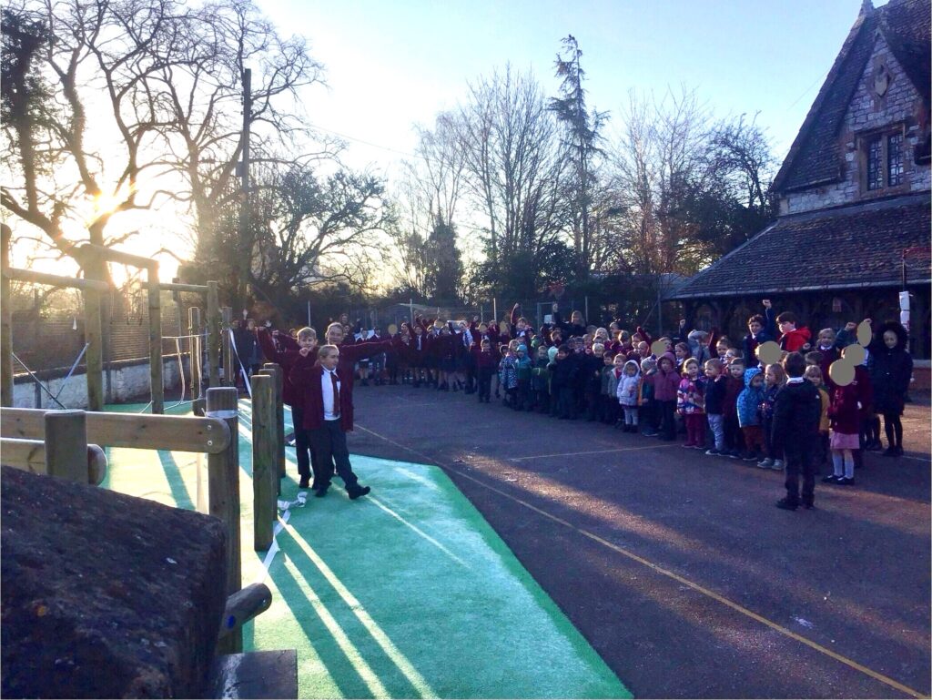 A group of children and adults stand in queues outside Lady Seawards Church of England Primary School on the playground in the early morning sunlight.
