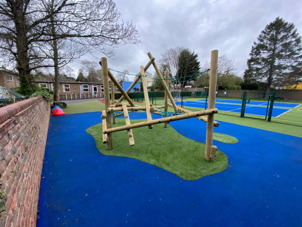 A wooden climbing frame stands on artificial grass and blue flooring beside tennis courts and a brick wall in an outdoor playground.