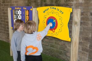Two children in grey hoodies interact with a colourful outdoor play clock mounted on a wall, whilst another maths activity board is visible nearby.