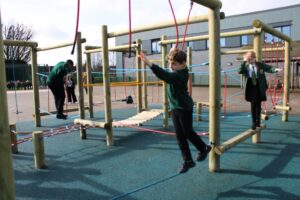 Three children in school uniforms play on a wooden climbing frame in an outdoor playground area with blue safety flooring.