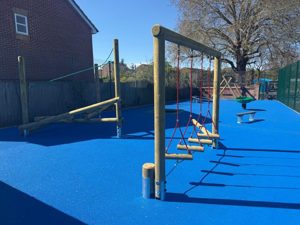 Playground with blue rubber flooring featuring wooden climbing structures, rope bridge, spinning seat, and a large tree in the background, bordered by a fence and brick building.