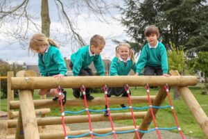 Four young children in school uniforms climb on a wooden playground structure with a rope net in an outdoor park.