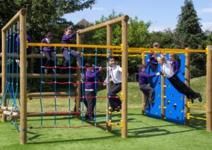 Children in school uniforms and an adult use a wooden and rope climbing frame and wall in an outdoor playground on a grassy area.