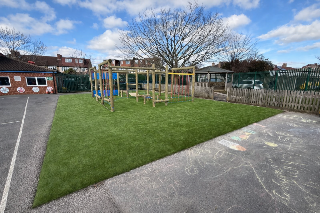 A playground with climbing equipment on artificial grass, bordered by asphalt with chalk drawings, near a school building and fenced area under a partly cloudy sky.